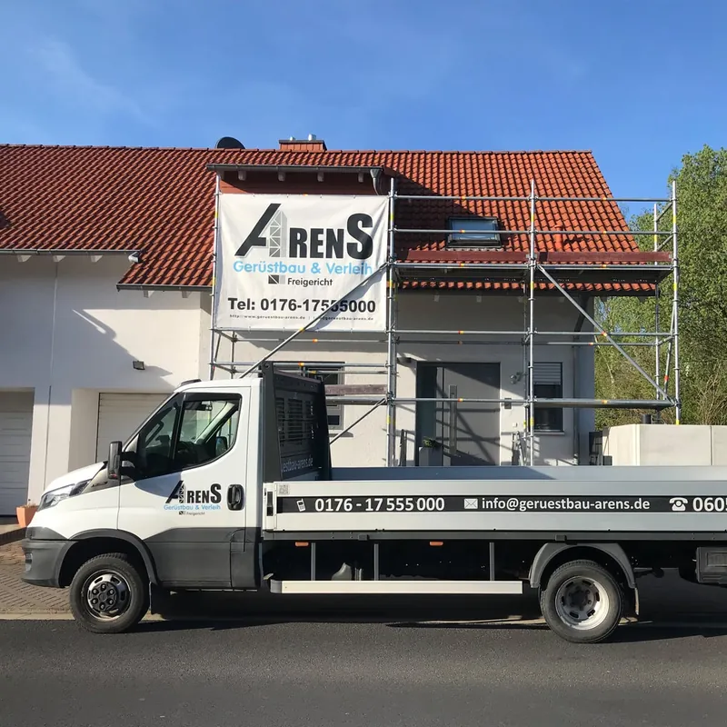 Weißer Arens-Pritschenwagen vor einem eingerüsteten Wohnhaus mit Arens-Banner