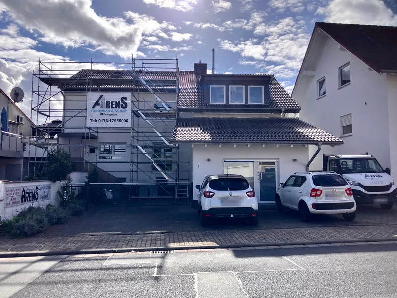 Eingerüstetes Wohnhaus mit Arens-Banner am Gerüst, davor zwei Autos und ein Arens-Transporter, an der Mauer ein Arens-Schild