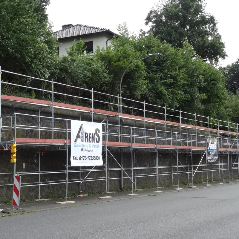 Langes Gerüst entlang einer hohen Mauer an einer Straße, mit Arens-Banner