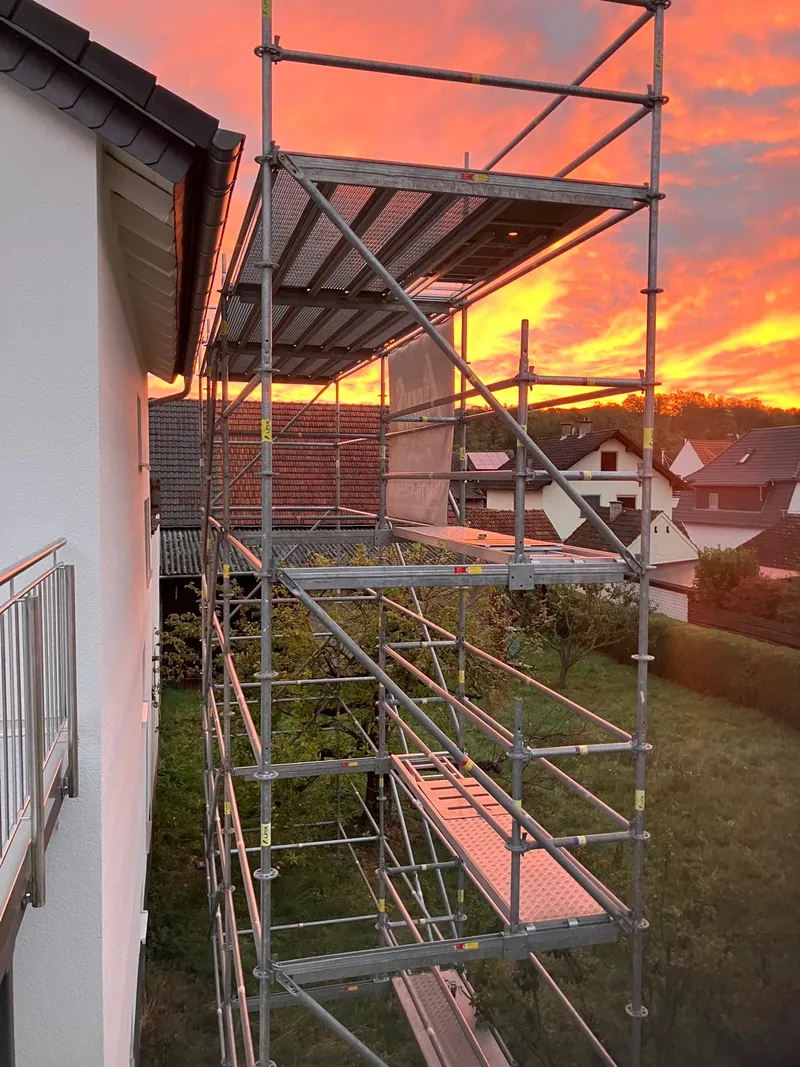 Gerüst mit Belägen an einer weißen Hauswand im Abendrot, unten Rasen
