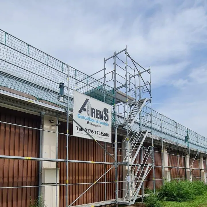Gerüst mit Treppenturm an einem langen Gebäude mit Holztoren, oben ein Arens-Banner