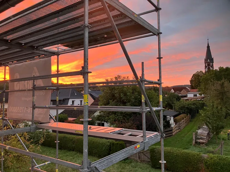 Gerüst mit Belägen an einem Wohnhaus im Abendrot, dahinter Gärten, Dächer und ein Kirchturm
