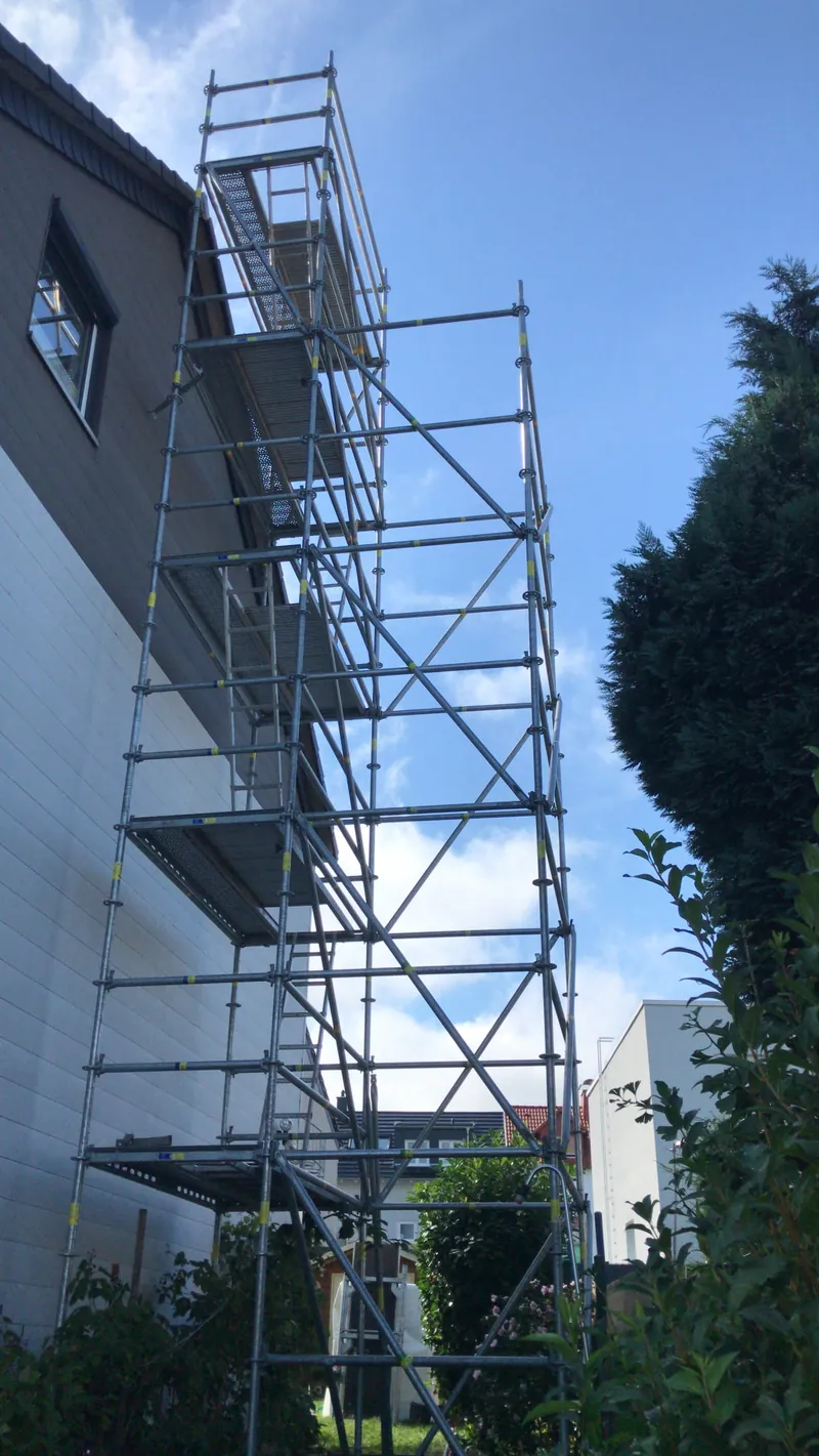 Schmaler, hoher Gerüstturm mit Treppenaufgang in einer engen Lücke neben einem Gebäude