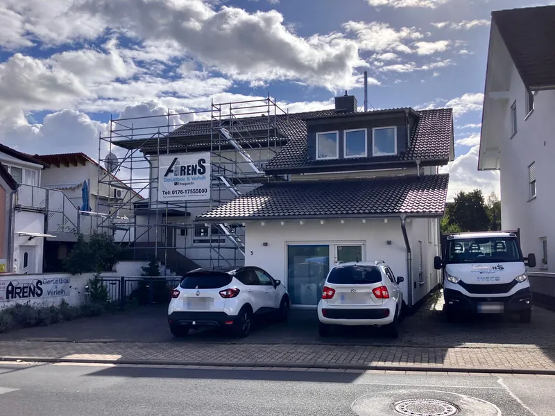 Wohnhaus mit Gerüst und Arens-Banner vor blauem Himmel, davor zwei Autos und ein Arens-Transporter
